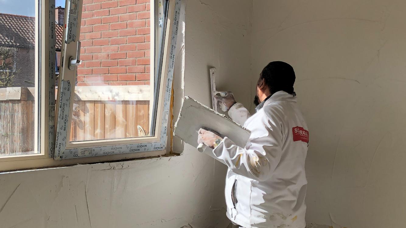 Sonny Bride stuct een wand naast een raam, in witte werkkleding met zijn rode logo