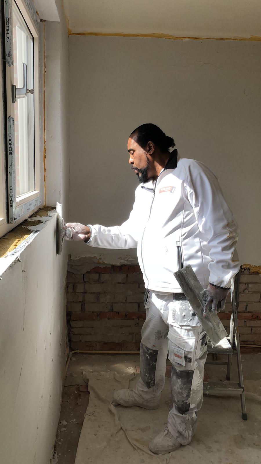 Sonny Bride strijkt een wand glad bij een raam, met spaan in de hand