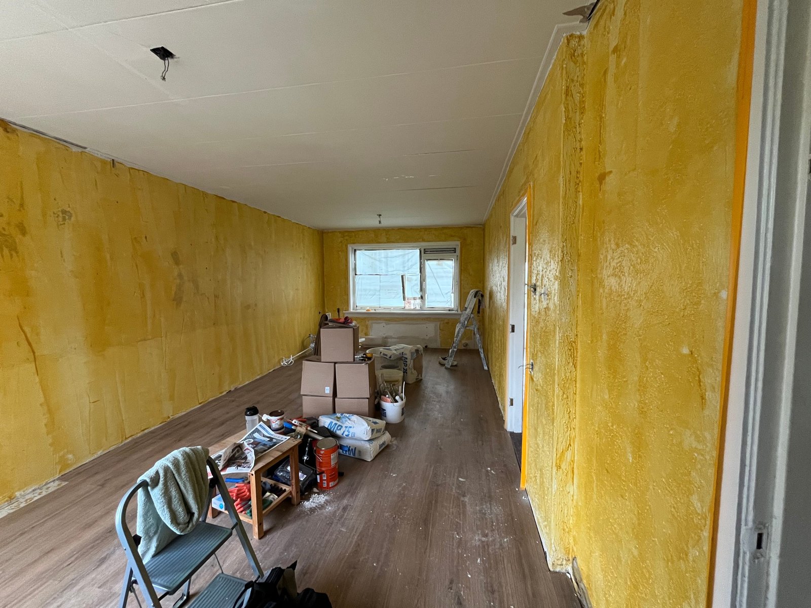 Woonkamer met oude gele wanden, ladder en verhuisdozen, voordat het stucwerk begint